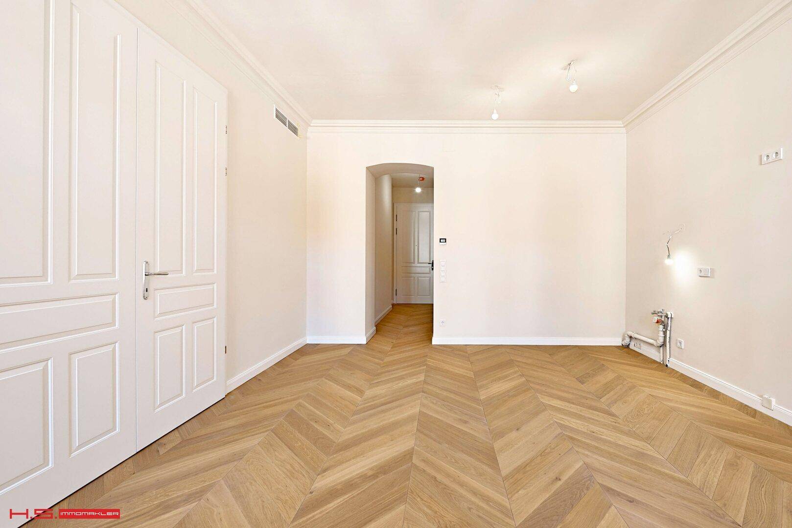 Leerer Raum mit Fischgrätparkett, hohen Decken und weiß gestrichenen Wänden.
