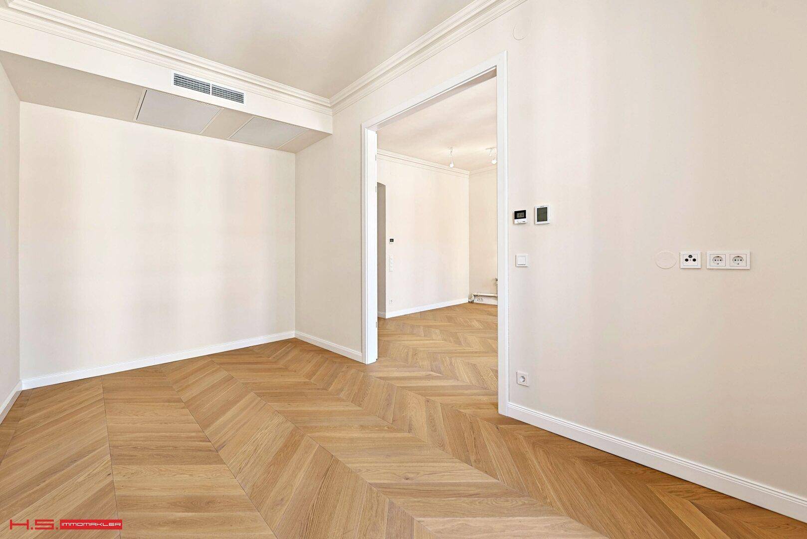 Heller Raum mit Fischgrätparkett und offenem Durchgang zu einem weiteren Zimmer.