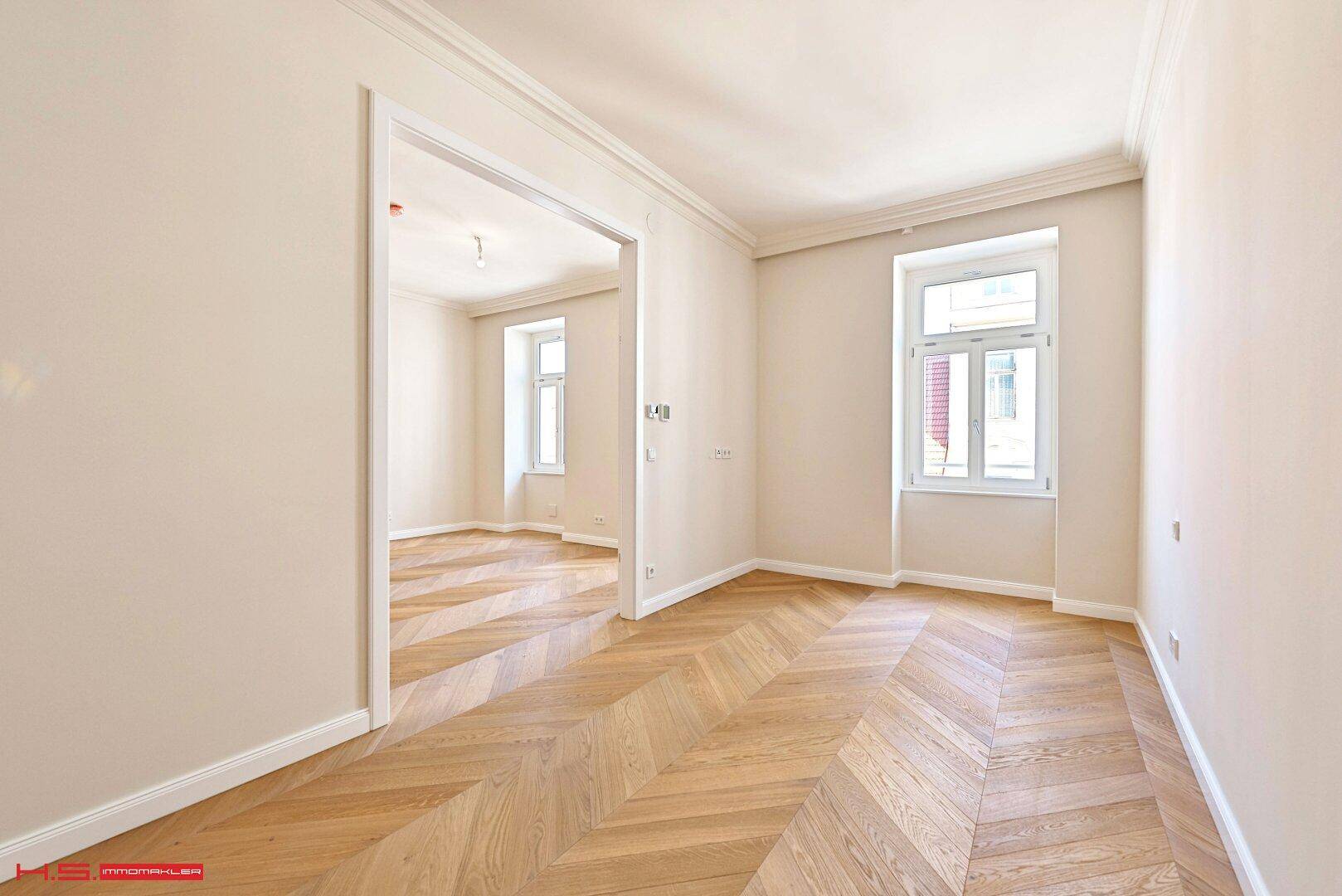 Heller Raum mit Fischgrätparkett und Blick durch eine Tür in ein weiteres Zimmer.