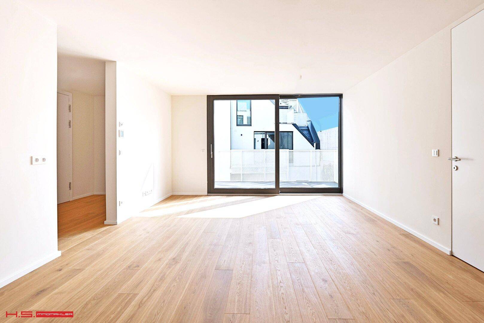 Leerer, heller Raum mit Holzboden und großer Fensterfront mit Blick auf den Innenhof.