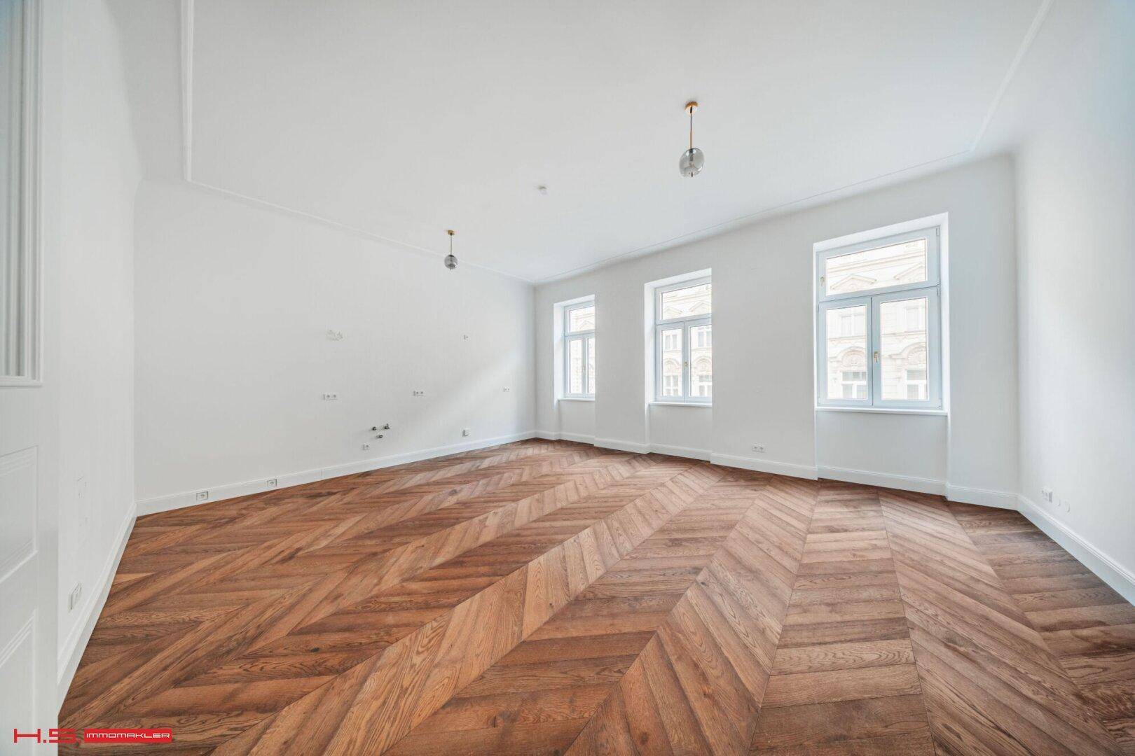 Leerer Raum mit Fischgrätparkett, hohen Decken und großen Fenstern für viel Tageslicht.