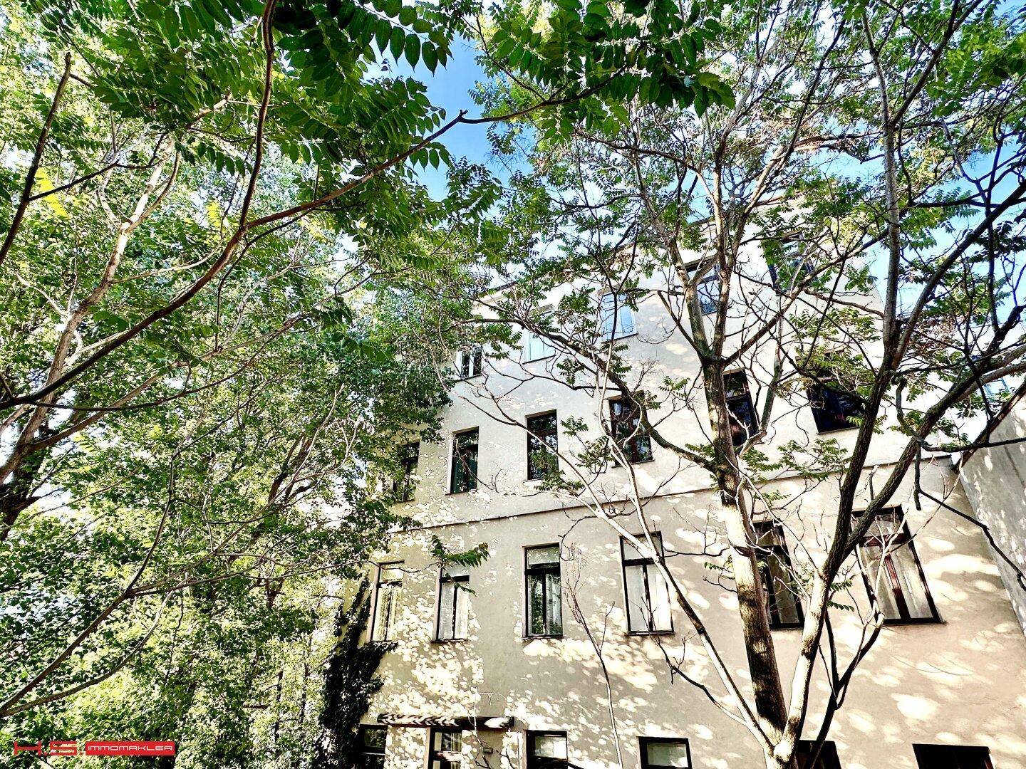 Fassade eines Wohngebäudes, umgeben von dichtem Grün und hohen Bäumen unter blauem Himmel.