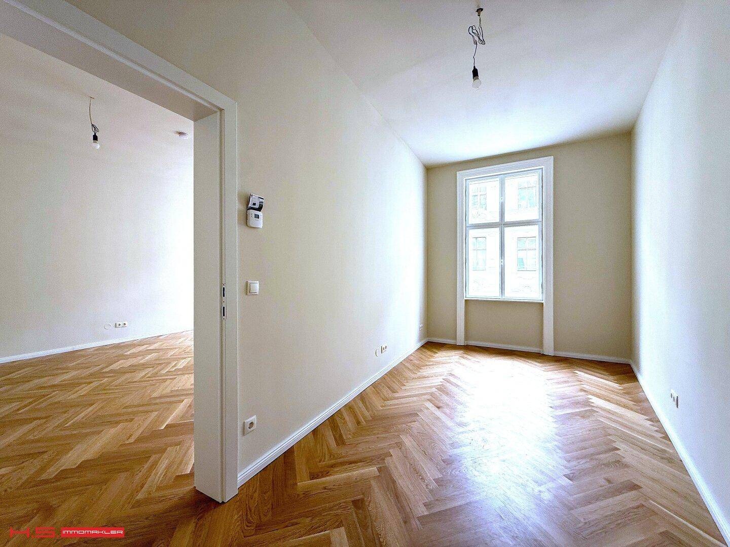 Heller Raum mit Fischgrätparkett und einem großen Fenster, das viel Licht hereinlässt.
