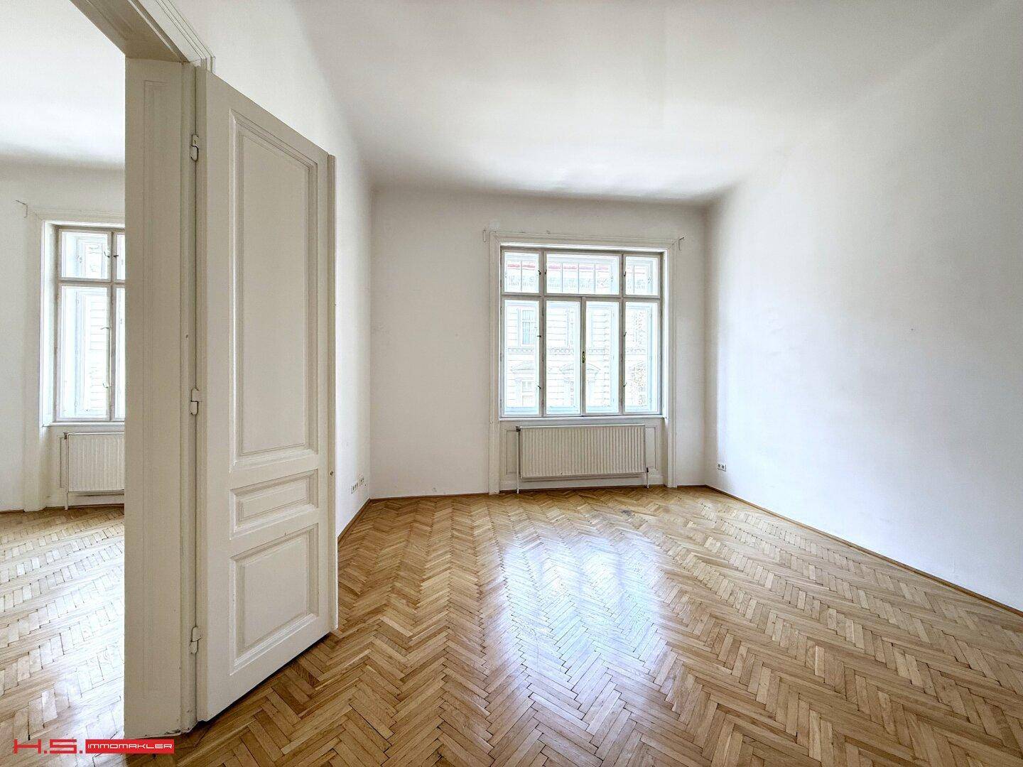 Geräumiges Zimmer mit Fischgrätparkett, hohen Decken und zwei großen Fenstern.