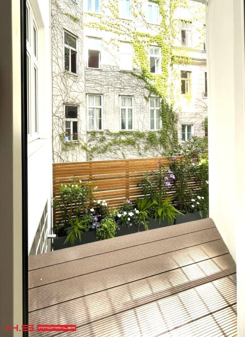 Kleiner Balkon mit Holzboden und Blick auf einen begrünten Innenhof mit Pflanzen.