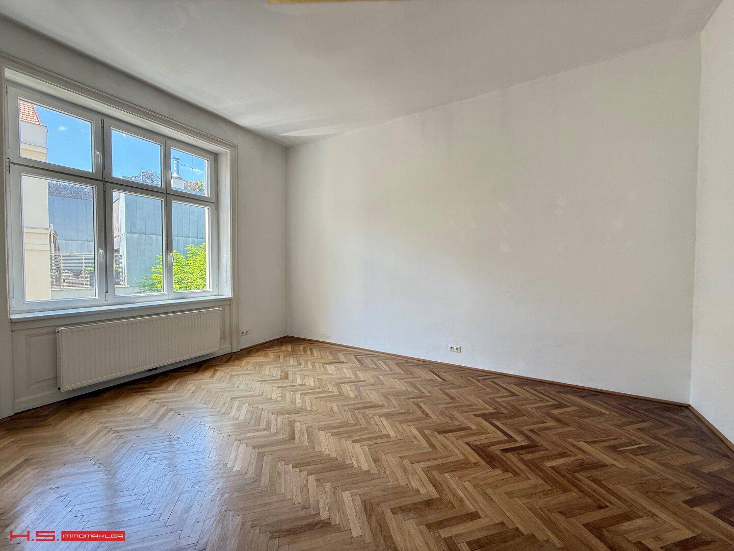 Geräumiges Zimmer mit klassischem Fischgrätparkett und großen Fenstern für eine helle Atmosphäre.