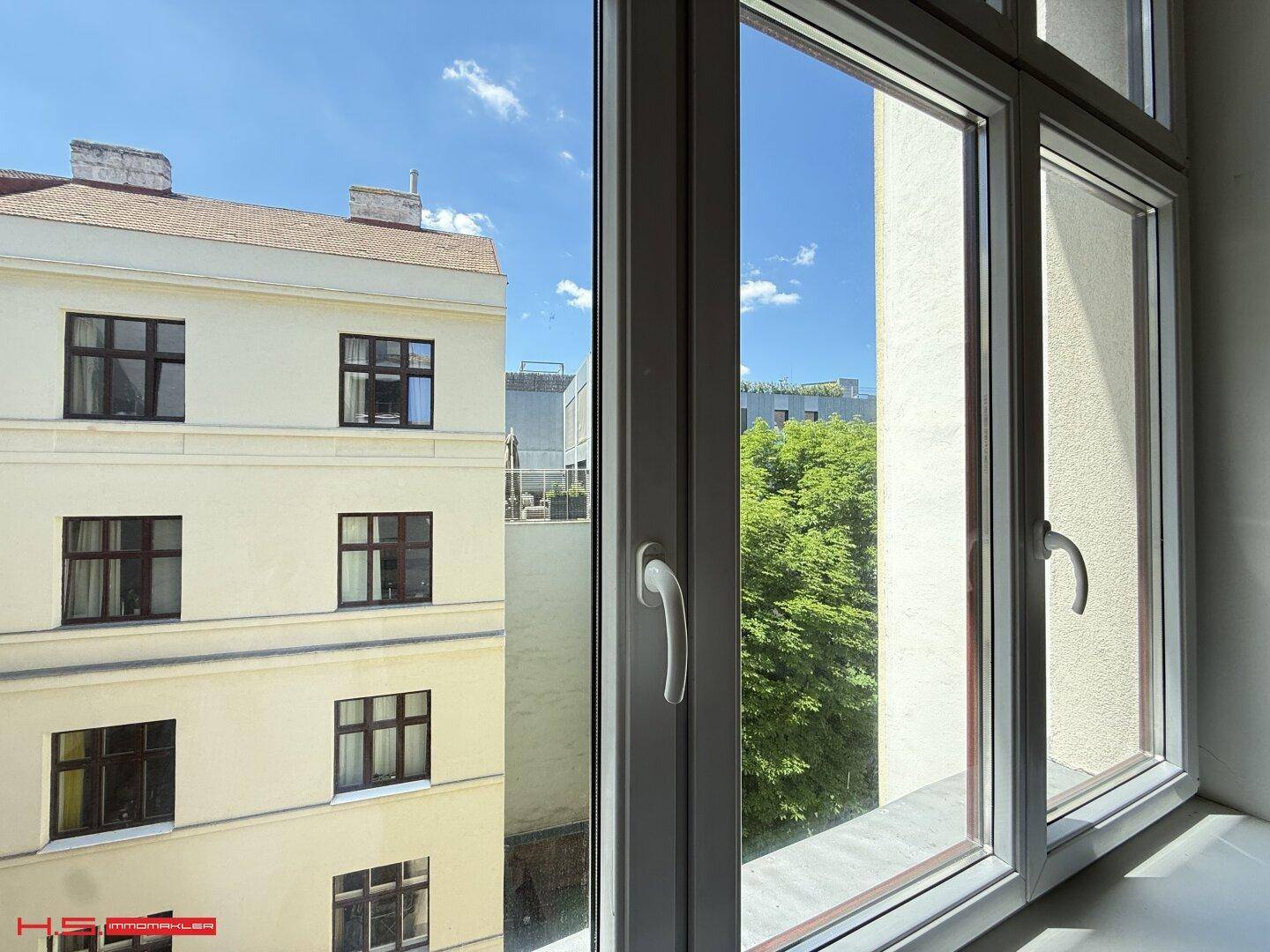 Blick aus dem Fenster auf ein Nachbargebäude und grüne Bäume unter blauem Himmel.