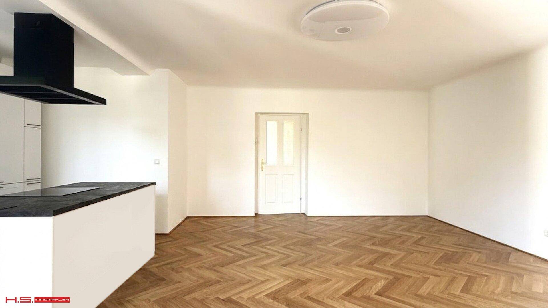 Heller Wohnbereich mit Fischgrätparkett und Zugang zu weiteren Räumen, angrenzend an die offene Küche.