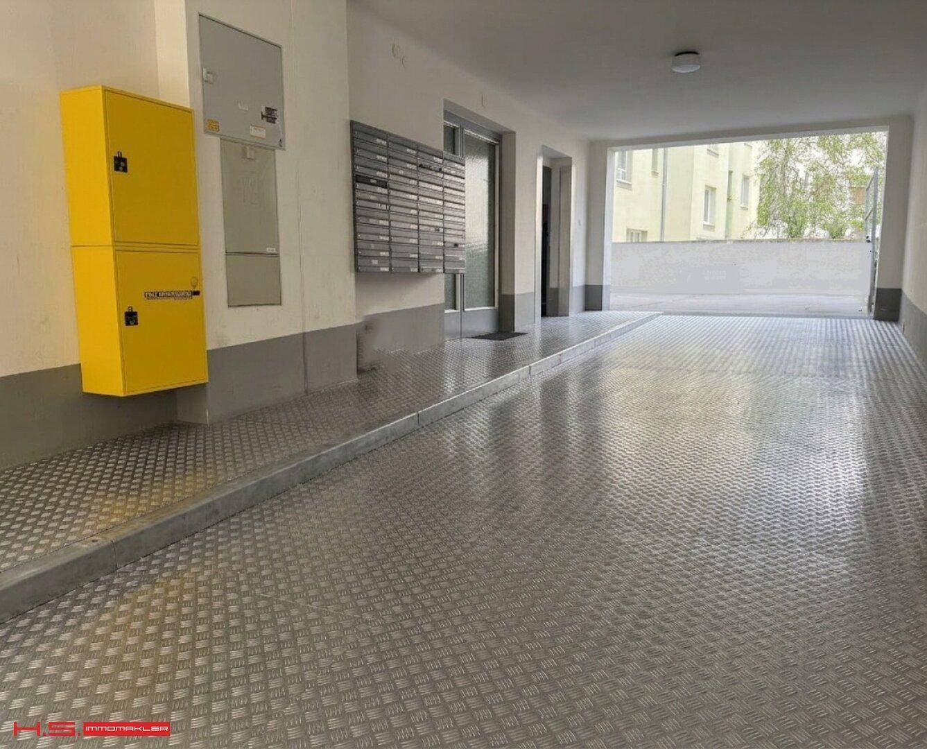 Eingangsbereich eines Gebäudes mit Metallboden, Briefkästen und Zugang zu einem Außenbereich.