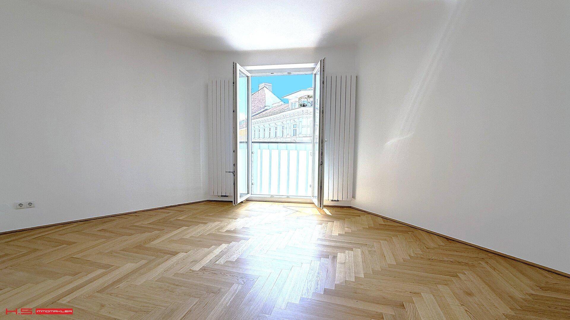 Lichtdurchflutetes Zimmer mit offenem französischen Balkon und Blick auf die umliegenden Gebäude.