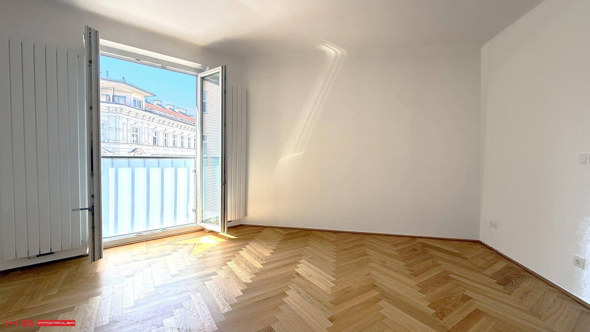Helles Zimmer mit Fischgrätparkettboden und einem französischen Balkon, der Sonnenlicht hereinlässt.