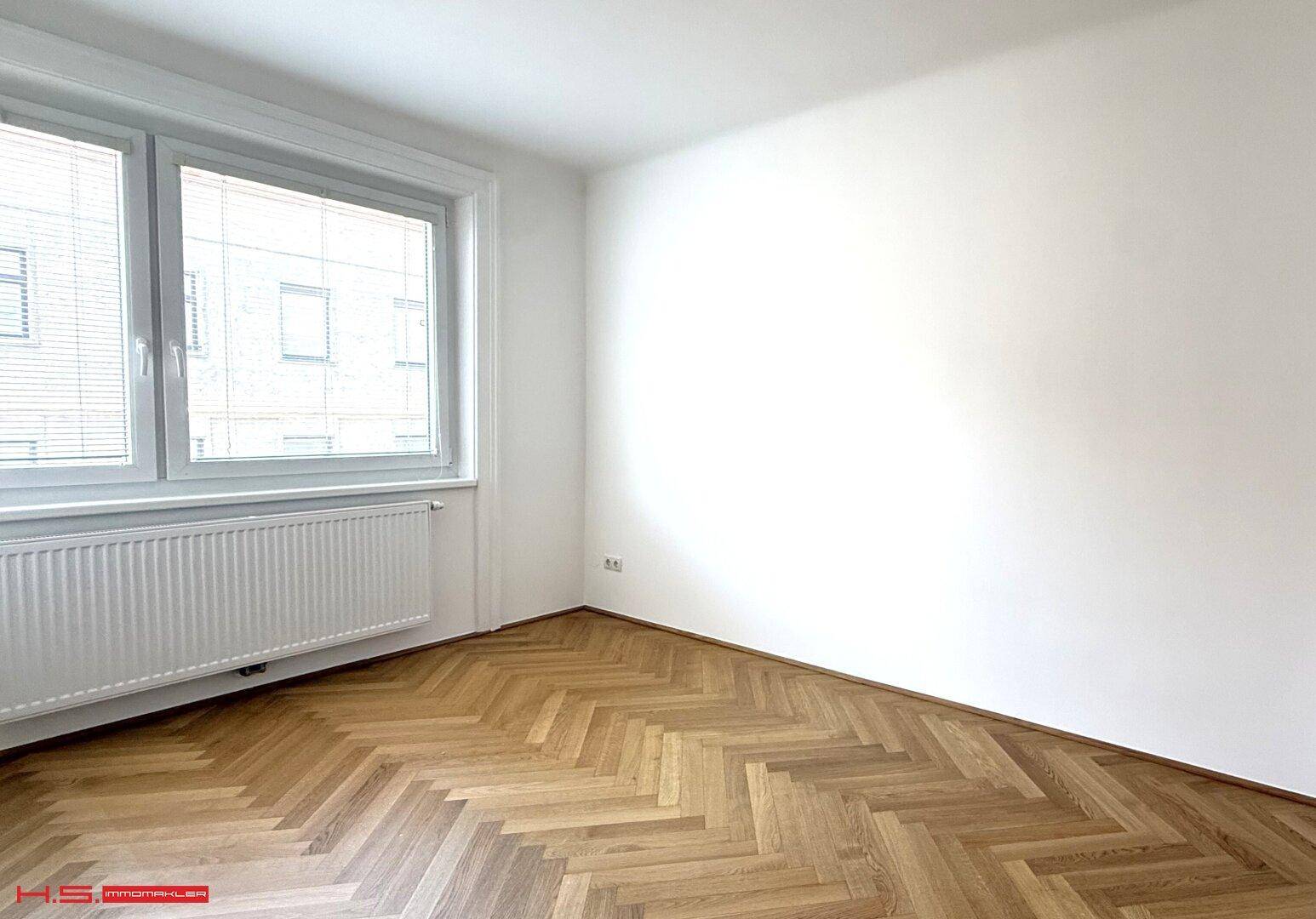 Leeres Zimmer mit Fischgrätparkettboden, weißen Wänden und einem Fenster mit Jalousien.