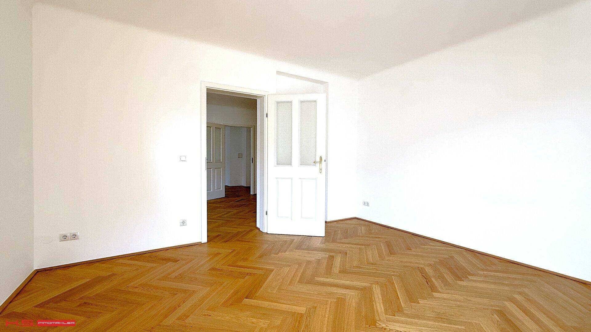 Helles Zimmer mit Fischgrätparkettboden und einer offenen Tür, die zu einem weiteren Raum führt.