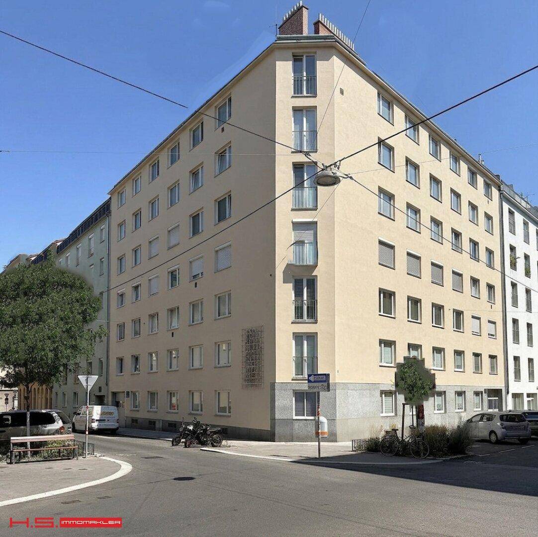 Mehrstöckiges Wohngebäude an einer Straßenecke mit heller Fassade und vielen Fenstern.