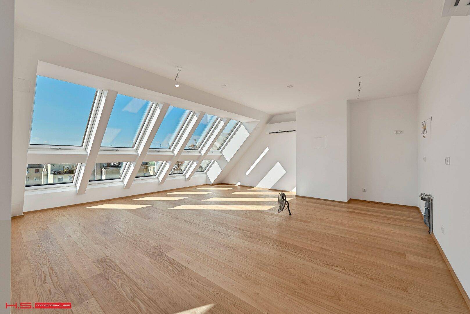 Heller, leerer Wohnraum mit Holzboden und großen Dachfenstern, die viel Licht hereinlassen.