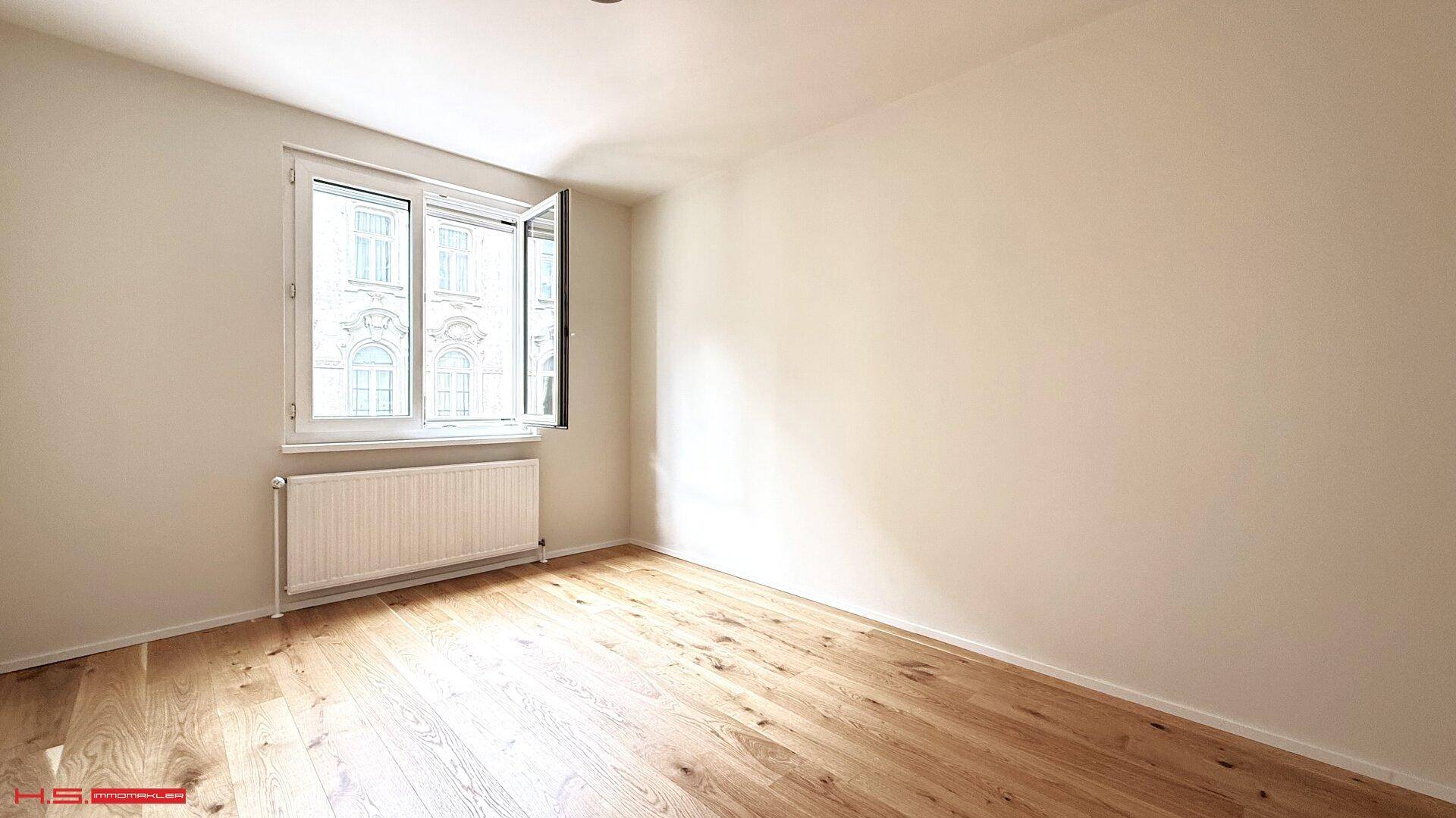 Helles Zimmer mit Holzboden und Fenster, das einen Blick auf die gegenüberliegende Architektur bietet.