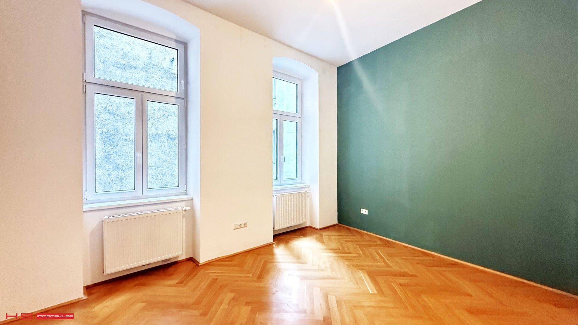 Helles Zimmer mit zwei Fenstern, Fischgrätparkett und einer farbigen Akzentwand.