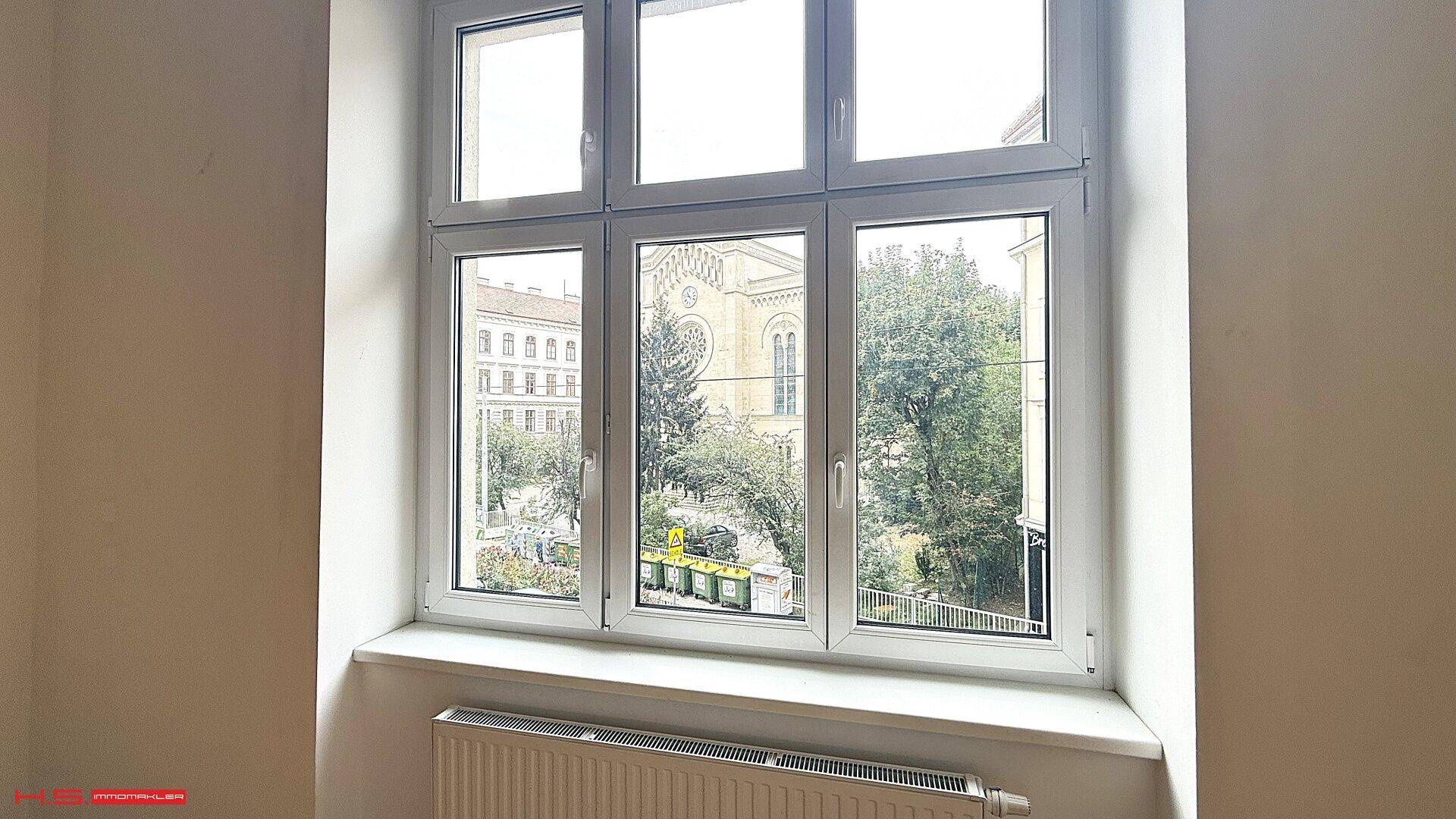Fensterblick auf eine Kirche und grüne Bäume, der viel Tageslicht hereinlässt.