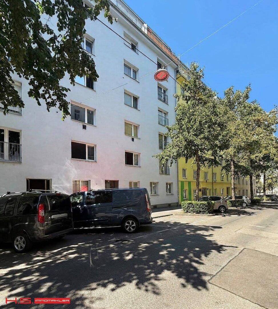 Wohngebäude mit weißer Fassade, vielen Fenstern und Bäumen davor an einem sonnigen Tag.