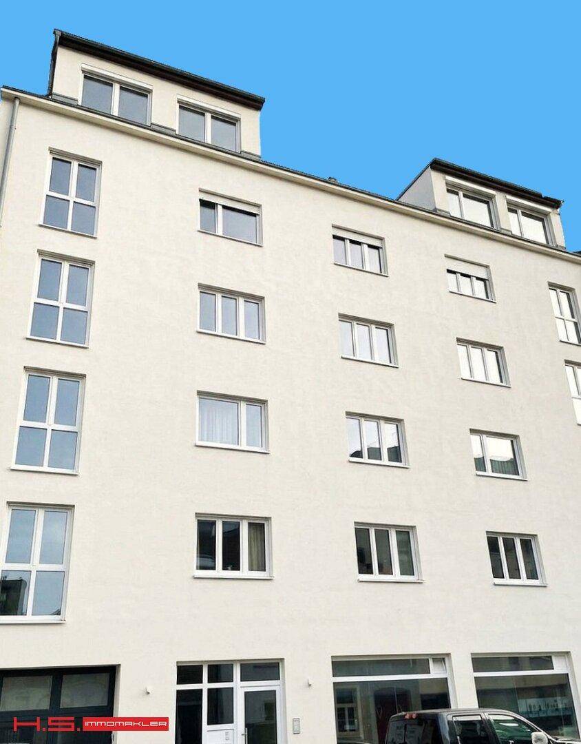 Modernes Mehrfamilienhaus mit heller Fassade, großen Fenstern und Dachgauben unter blauem Himmel.