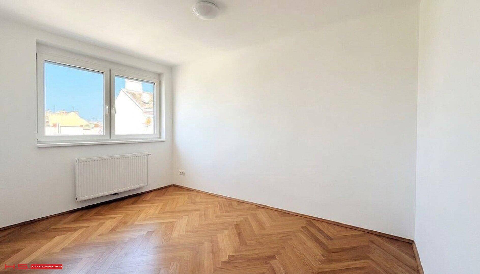 Helles Zimmer mit elegantem Fischgrätparkettboden, großen Fenstern und weiß gestrichenen Wänden.