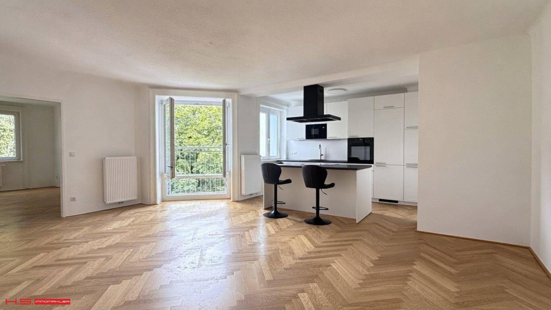 Heller Wohn- und Essbereich mit Fischgrätparkett, offener Küche und Zugang zu einem französischen Balkon.