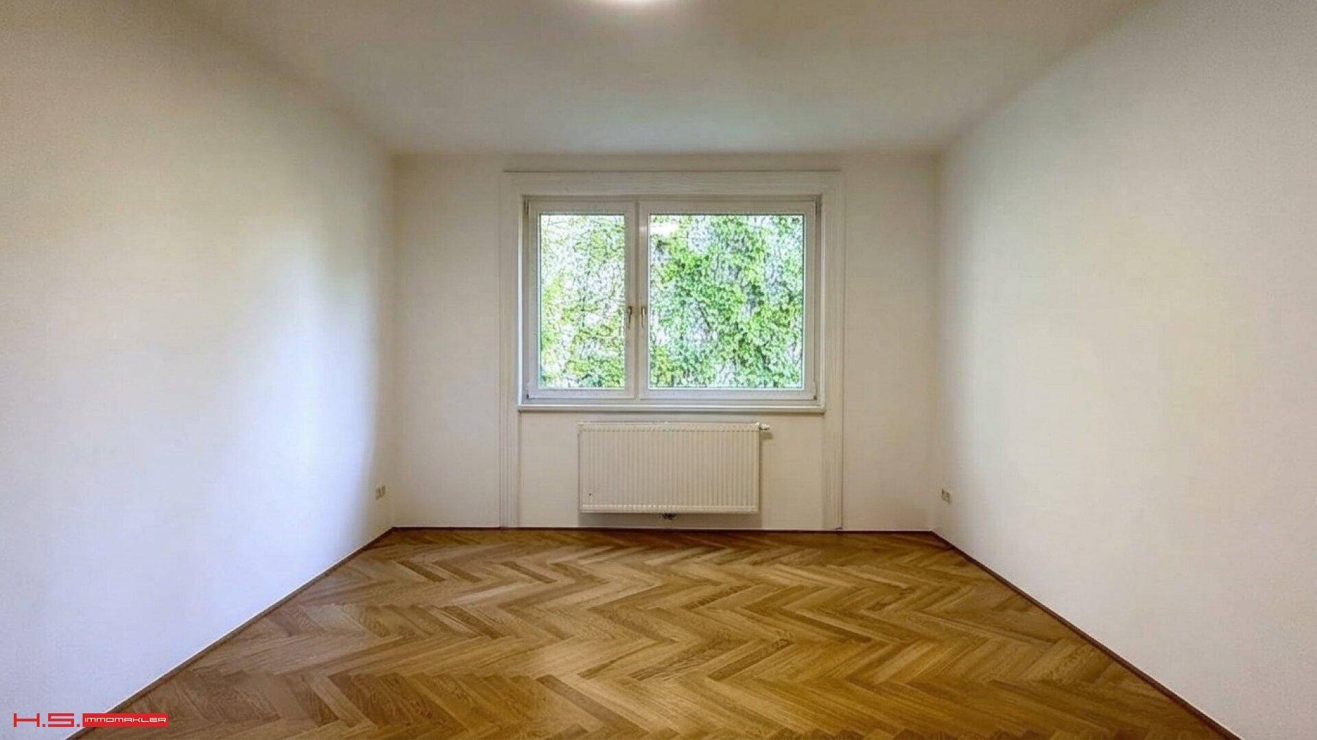 Geräumiges Zimmer mit Fischgrätparkett und Fenster mit Blick auf üppiges Grün.