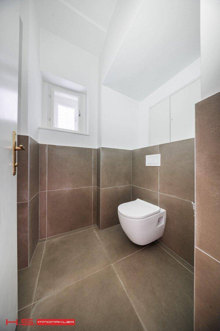 Separates WC mit wandhängender Toilette, kleinem Fenster und modernen Fliesen.