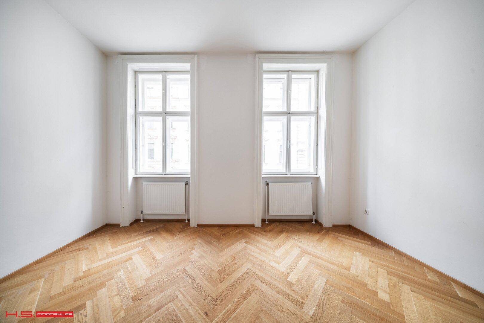 Lichtdurchflutetes Zimmer mit zwei großen Fenstern und elegantem Fischgrätparkettboden.