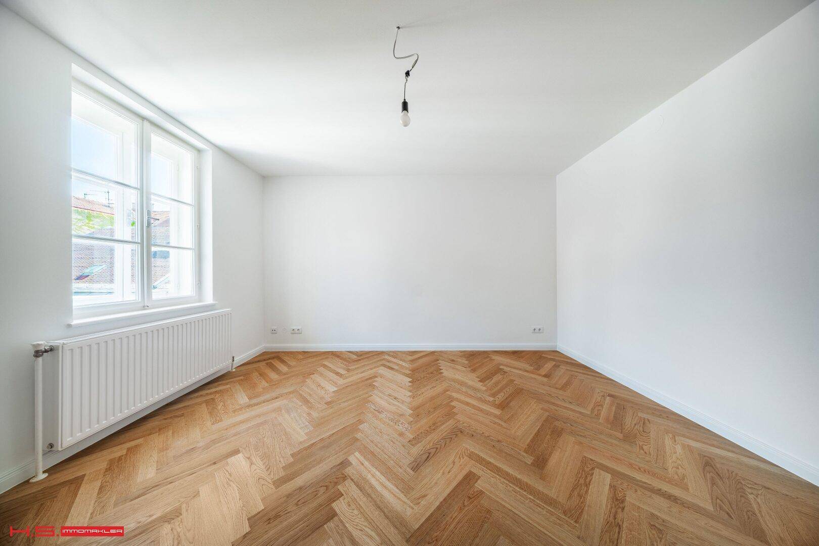Geräumiges Zimmer mit hellem Fischgrätparkett und großen Fenstern, die viel Licht hereinlassen.