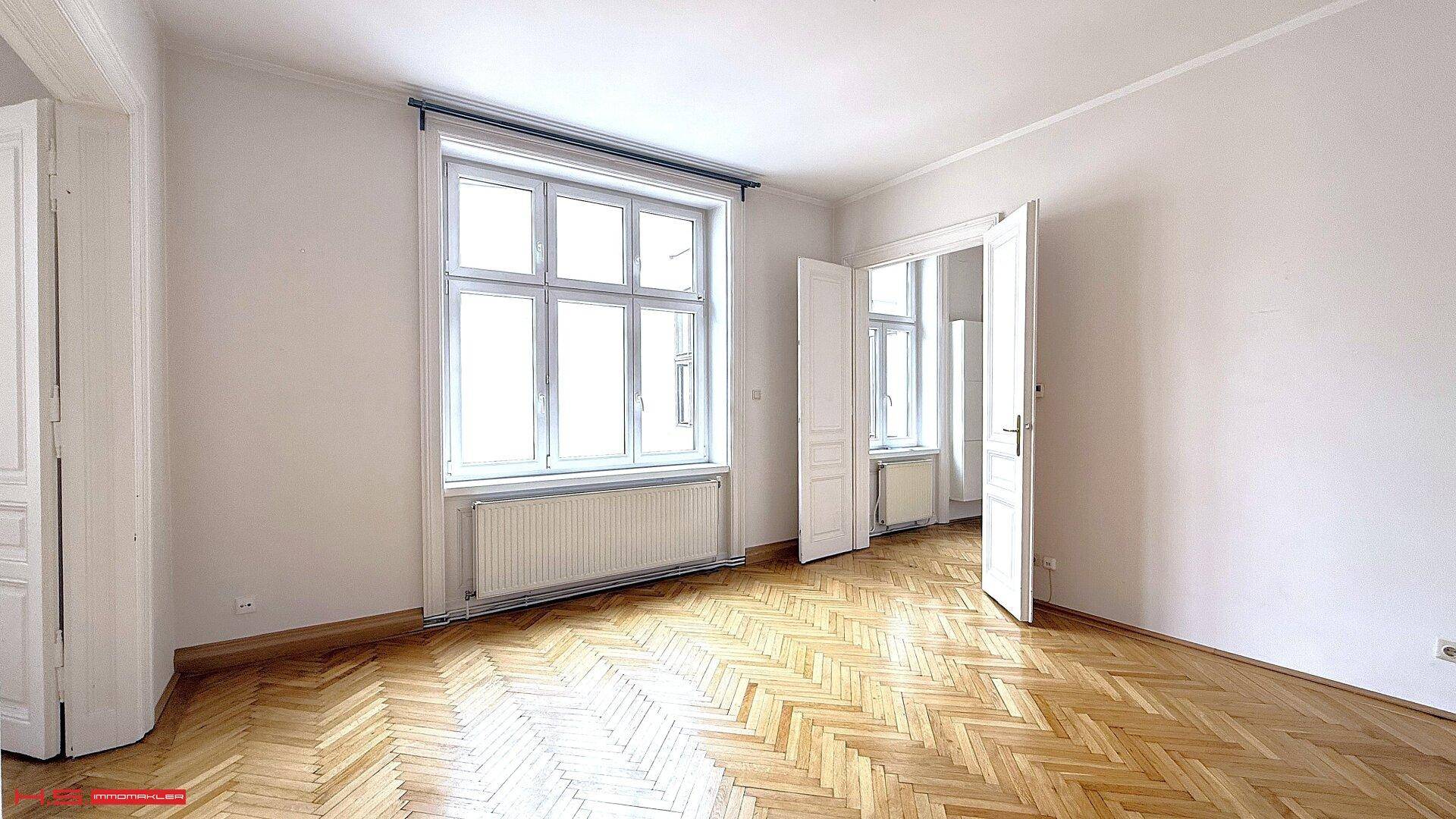 Geräumiges Zimmer mit Fischgrätparkett und großen Fenstern, das viel Licht hereinlässt.