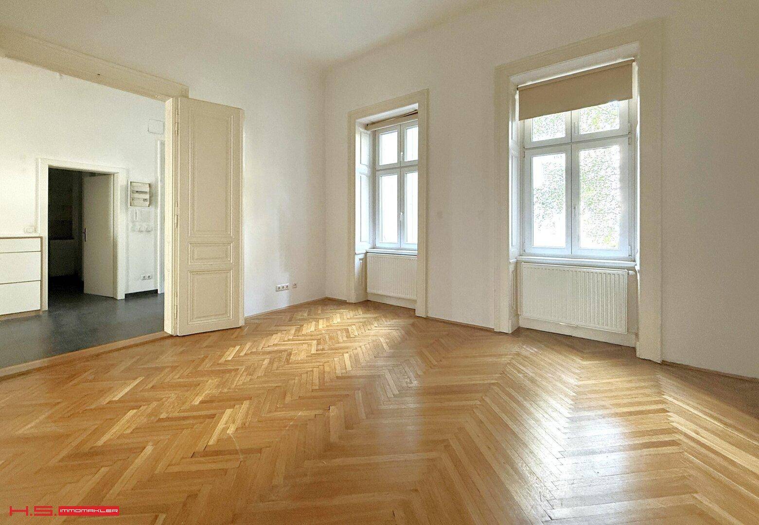 Geräumiges Zimmer mit Fischgrätparkett und zwei großen Fenstern, die viel Sonnenlicht hereinlassen.