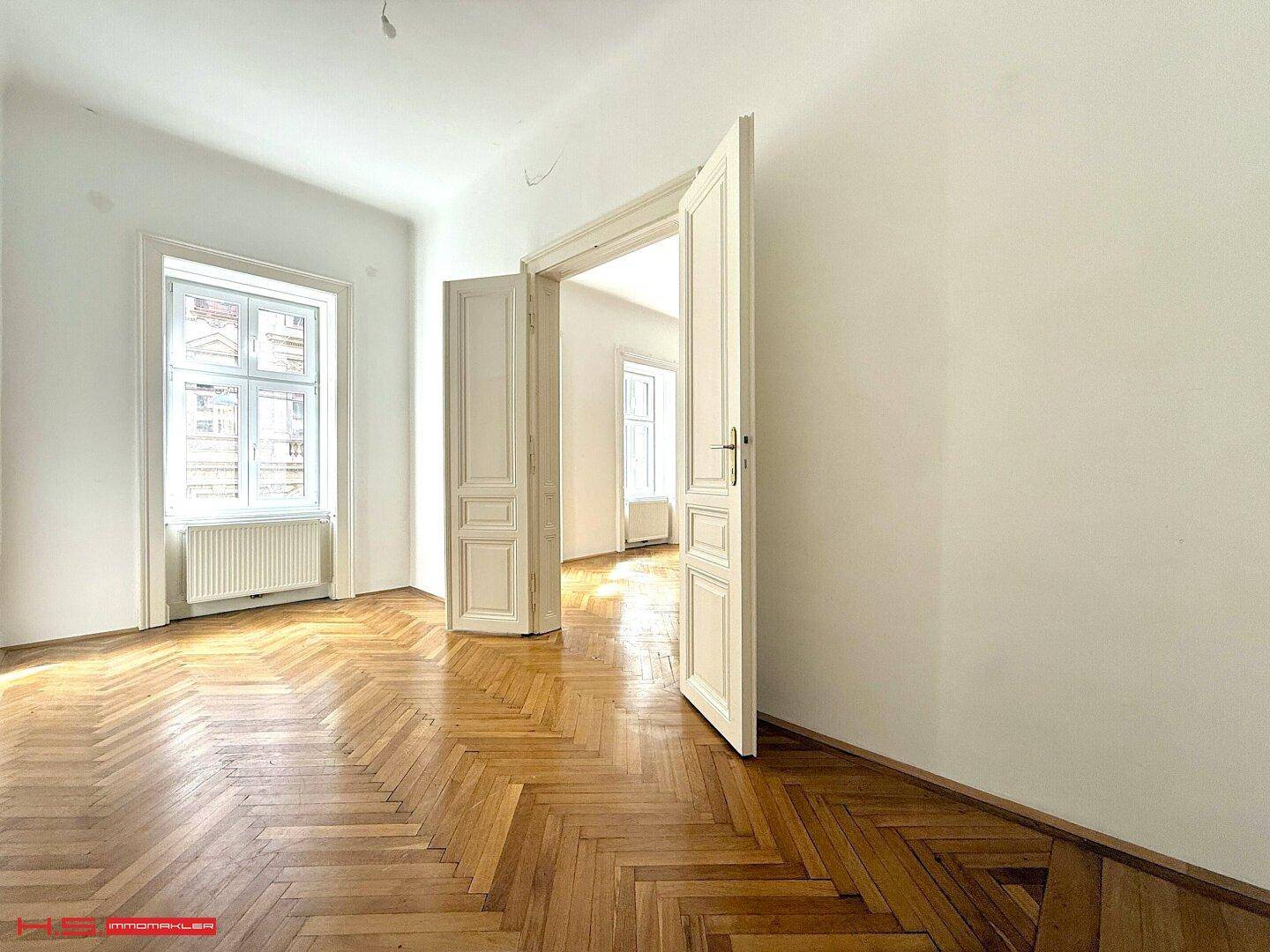 Geräumiges Zimmer mit klassischem Fischgrätparkett und hohen Doppeltüren zu einem weiteren hellen Raum.