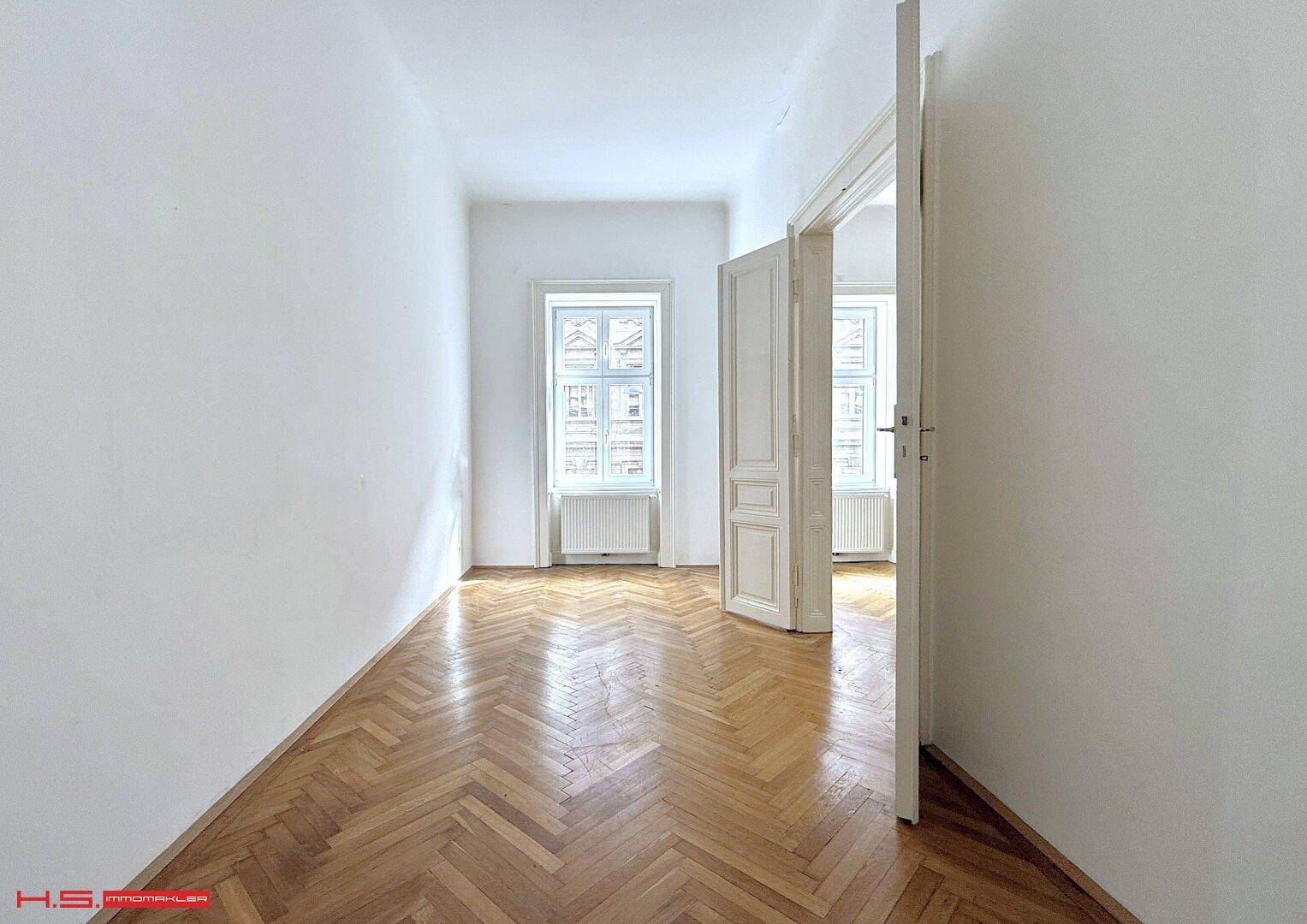 Leeres Zimmer mit Fischgrätparkett und hohen Türen, die zu einem weiteren hellen Raum führen.