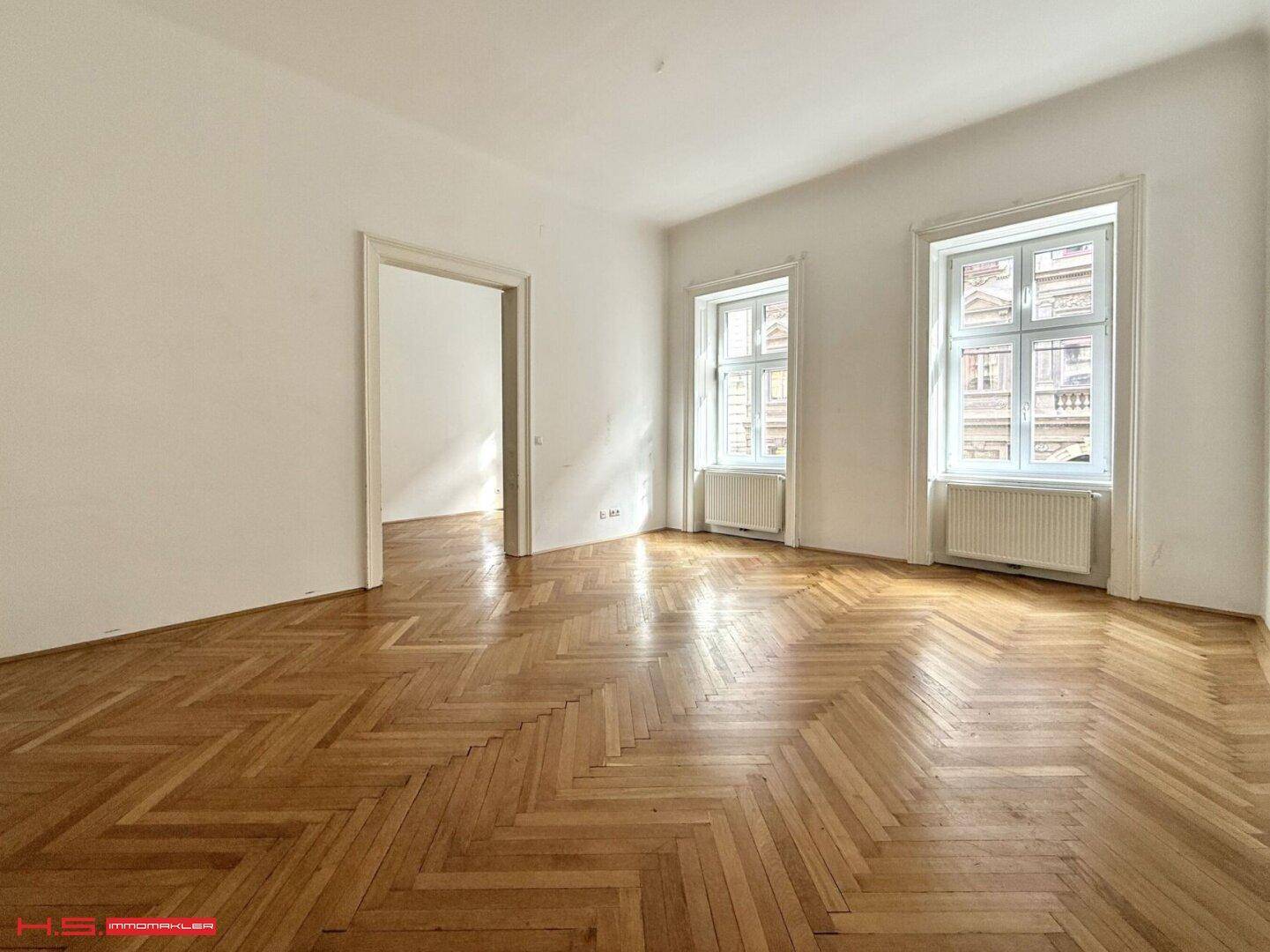 Heller Raum mit Fischgrätparkett und zwei großen Fenstern, die den Blick nach draußen freigeben.
