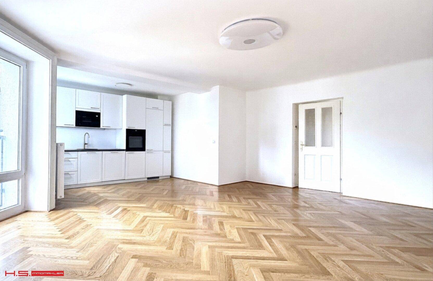Moderne Einbauküche mit weißen Schränken, dunkler Arbeitsplatte und Fischgrätparkett in einem offenen Raum.
