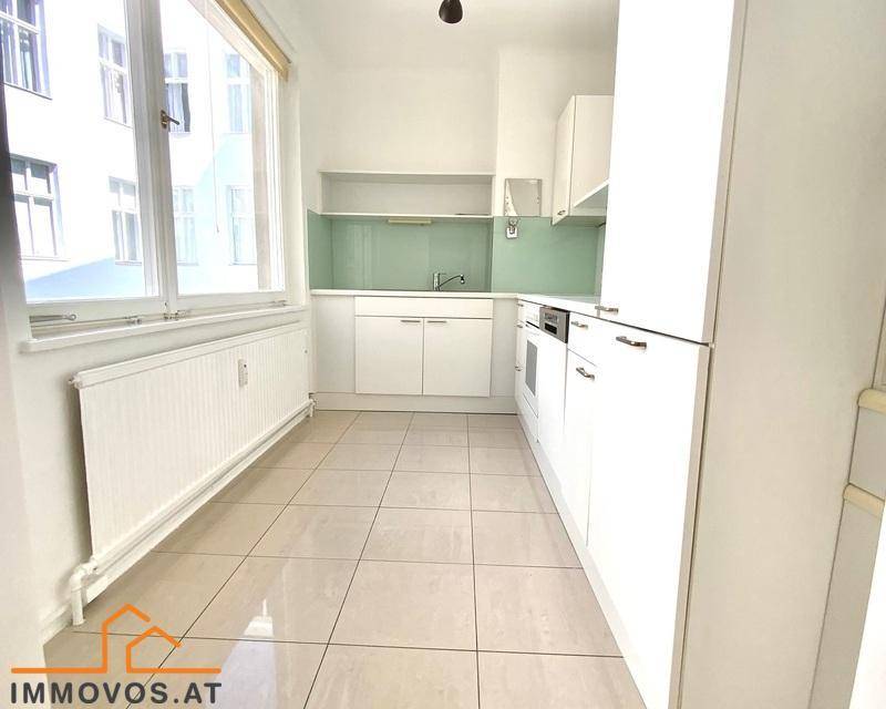 Funktionale Küche mit weißen Schränken, Fliesenboden und einem großen Fenster für ausreichend Tageslicht.