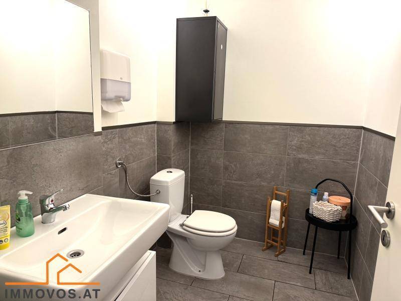 Modernes WC mit grauen Fliesen, wandmontierter Toilette und einem Waschbecken mit Unterschrank.