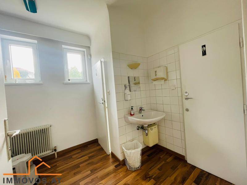 Helle Toilette mit Waschbecken, Fliesen an der Wand und zwei Fenstern für natürliche Belüftung.