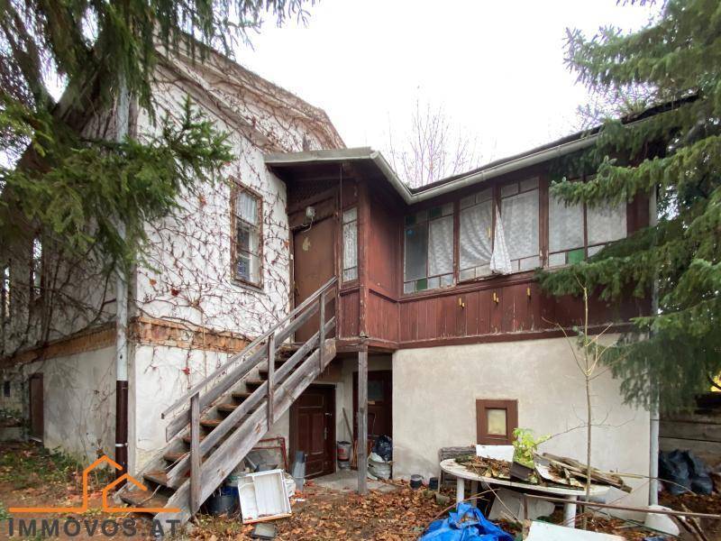 Rückseite des Gebäudes mit einer Holztreppe, Verkleidung und starkem Pflanzenbewuchs.