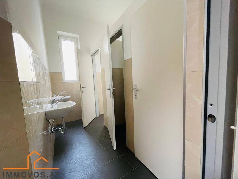 Waschraum mit mehreren Toilettenkabinen, zwei Waschbecken, einem Fenster und Fliesen an Wänden und Boden.