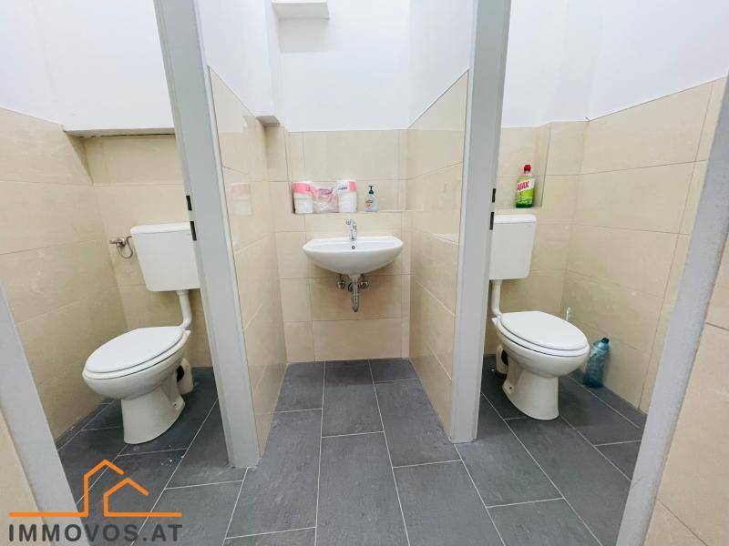Öffentliche Toilettenanlage mit zwei Kabinen, einem gemeinsamen Waschbecken und Fliesenboden.