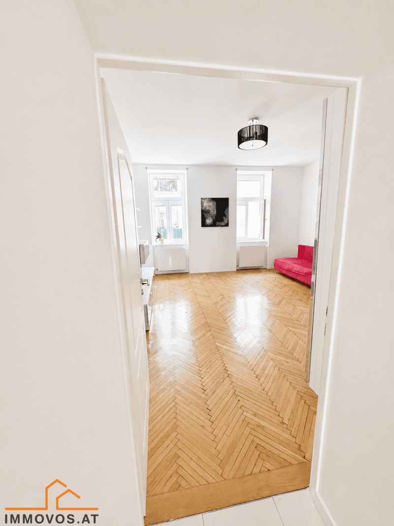Blick durch eine Tür in das helle Wohnzimmer mit durchgehendem Fischgrätparkett und großen Fenstern.