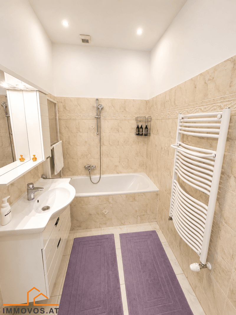Badezimmer mit beigefarbenen Fliesen, Badewanne mit Duschfunktion und beheiztem Handtuchhalter.