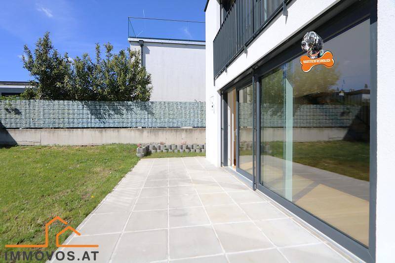 Gepflasterte Terrasse neben einer Rasenfläche mit großen Fenstern zum Haus.