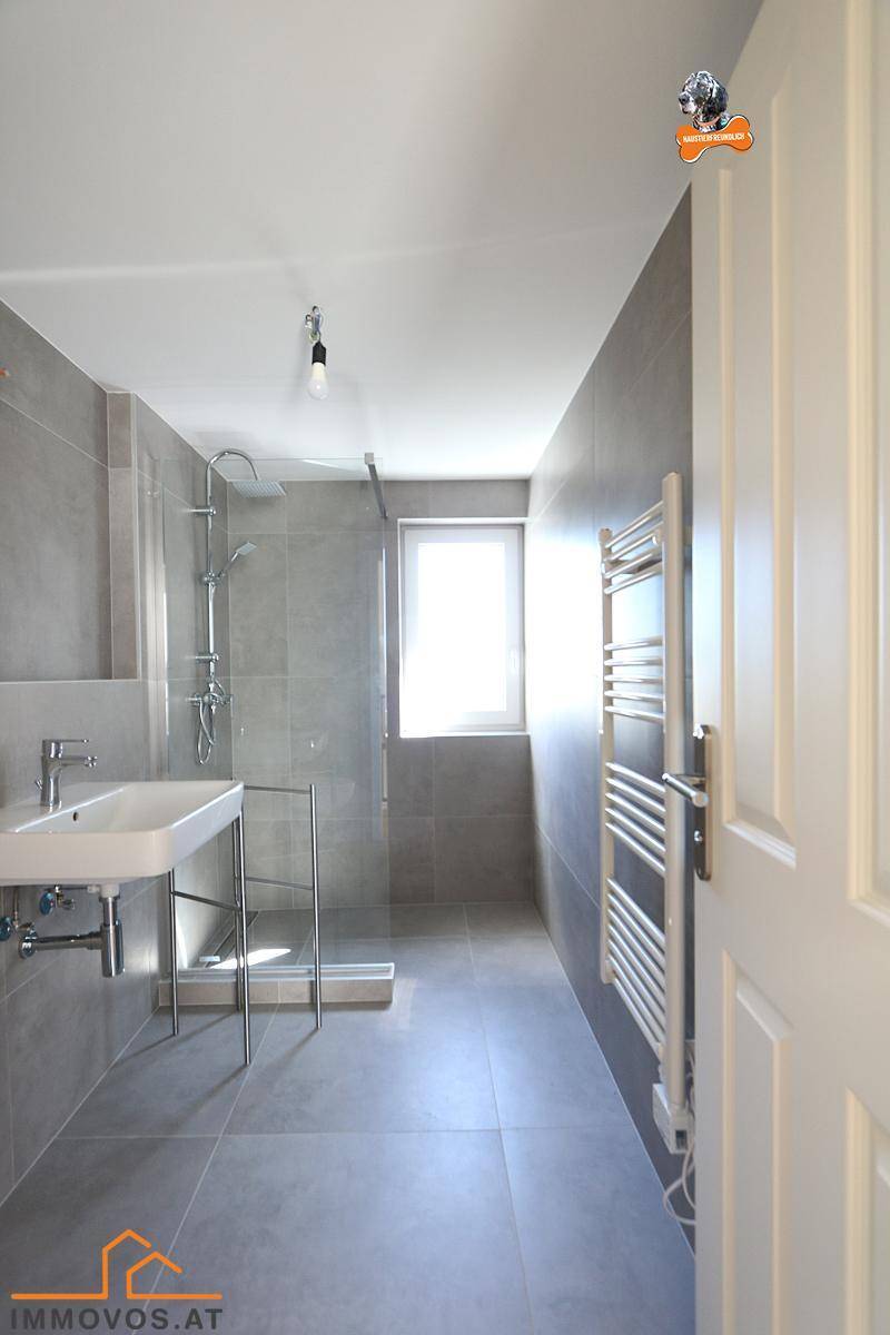 Helles Badezimmer mit Fenster, grauen Fliesen, Dusche und beheiztem Handtuchhalter.