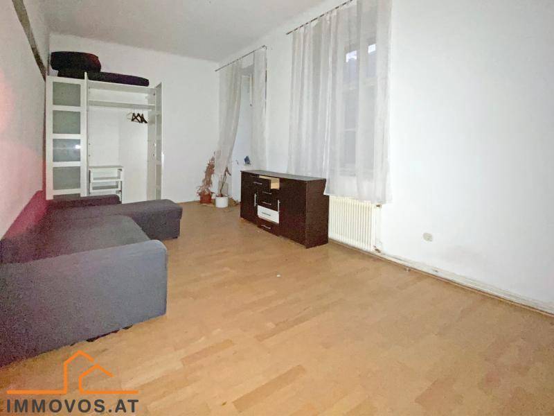 Helles Wohnzimmer mit Parkettboden, großem Fenster und einfacher Einrichtung, bietet viel Potenzial.