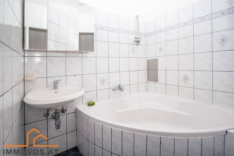 Badezimmer mit Eckbadewanne, Waschbecken und weißen Fliesen, in gutem, aber älterem Zustand.