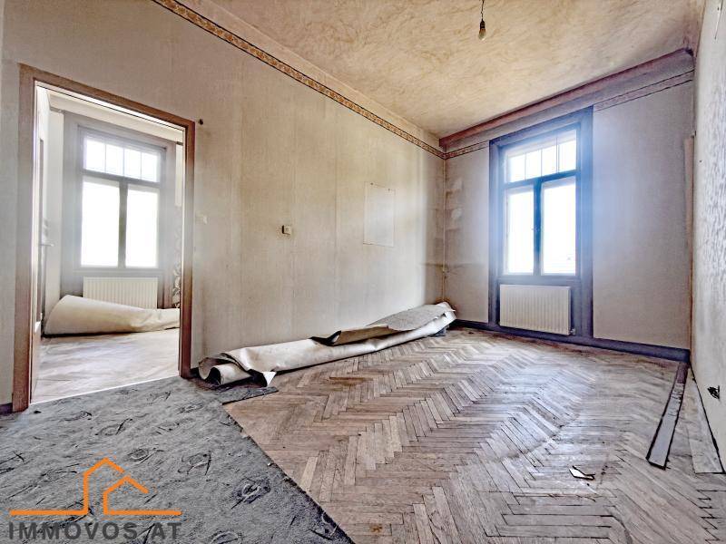 Zimmer mit altem Fischgrätparkett, hohen Decken und Blick in einen weiteren Raum.