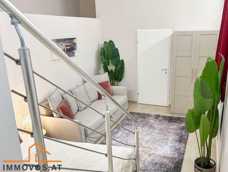 Gemütlicher Wohnbereich mit Sofa, Teppich und Pflanzen, verbunden durch eine moderne Treppe zu einer oberen Ebene.